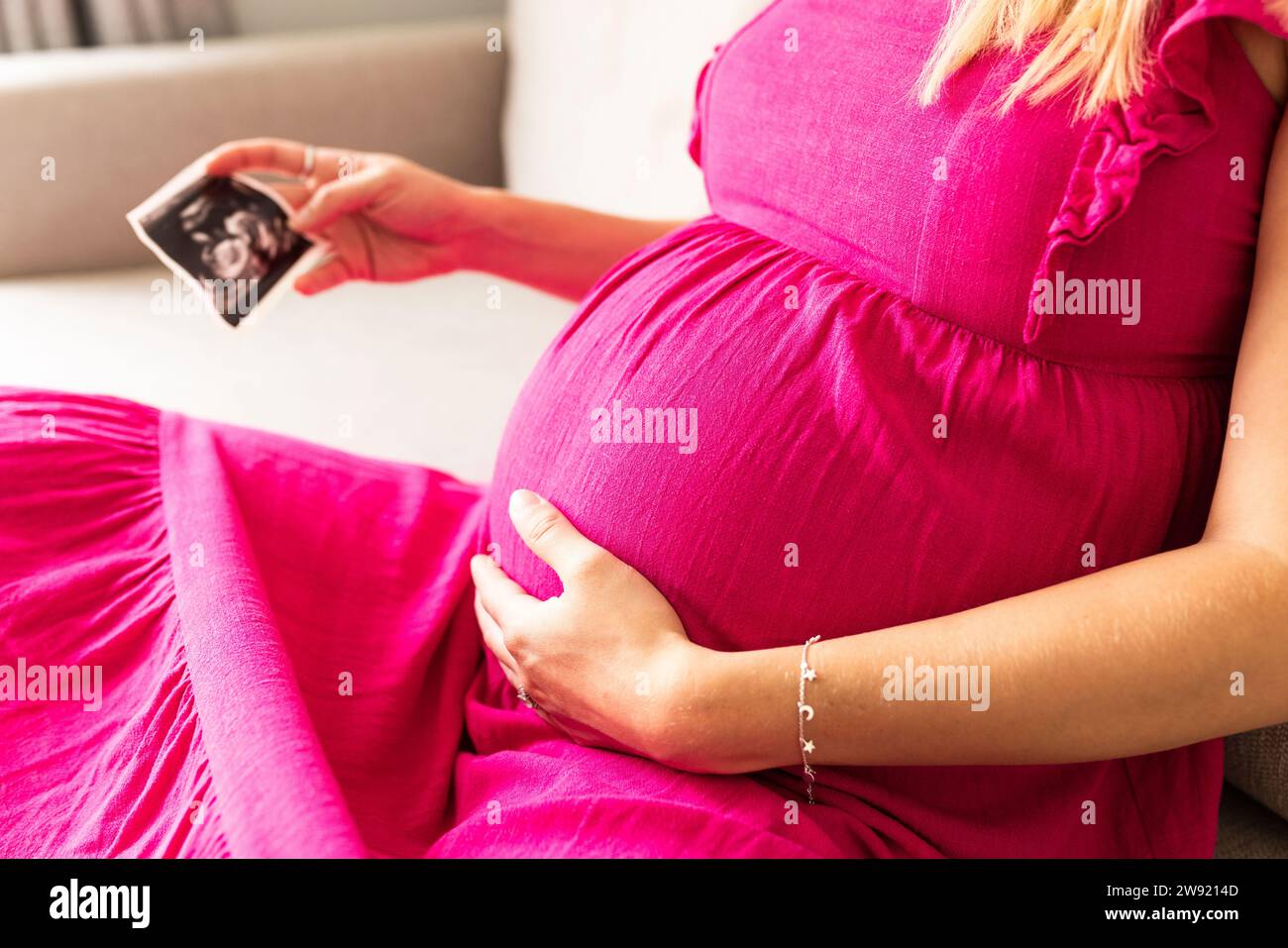Schwangere Frau, die zu Hause ein Ultraschallfoto mit der Hand auf dem Magen hält Stockfoto