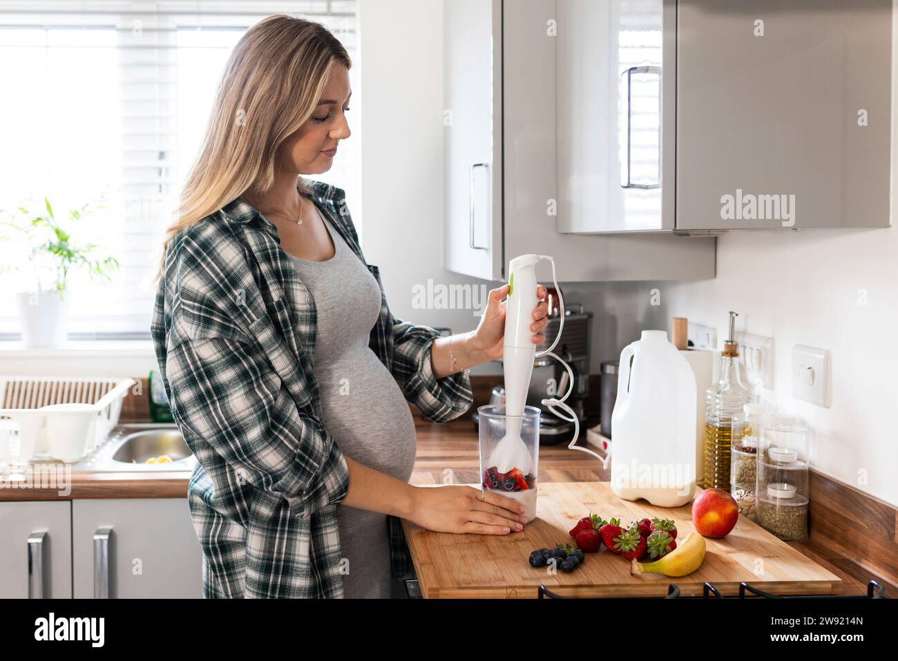 Blonde schwangere Frau, die Milchshake mit Stabmixer in der Küche zubereitet Stockfoto
