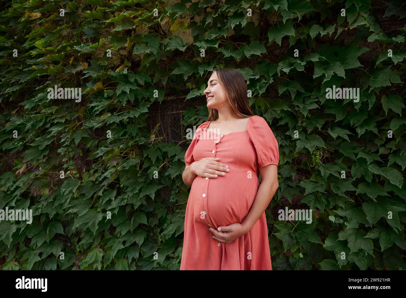 Glückliche schwangere Frau, die Kleid trägt und vor Pflanzen steht Stockfoto