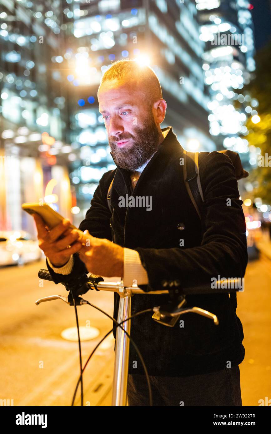 Mann, der sein Smartphone benutzt und sich nachts in der Stadt auf den Radweg gestützt hat Stockfoto