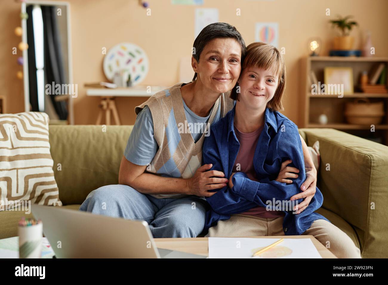 Lächelnde Mutter und ihre Tochter mit Down-Syndrom sitzen auf dem Sofa Stockfoto