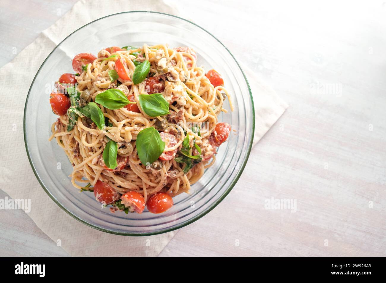 Kalter Spaghetti-Salat mit Tomaten, Rucola, Mozzarella, Oliven und einem cremigen Dressing mit Basilikumgarnitur in einer Glasschale auf einem weiß bemalten Tisch, oben Stockfoto