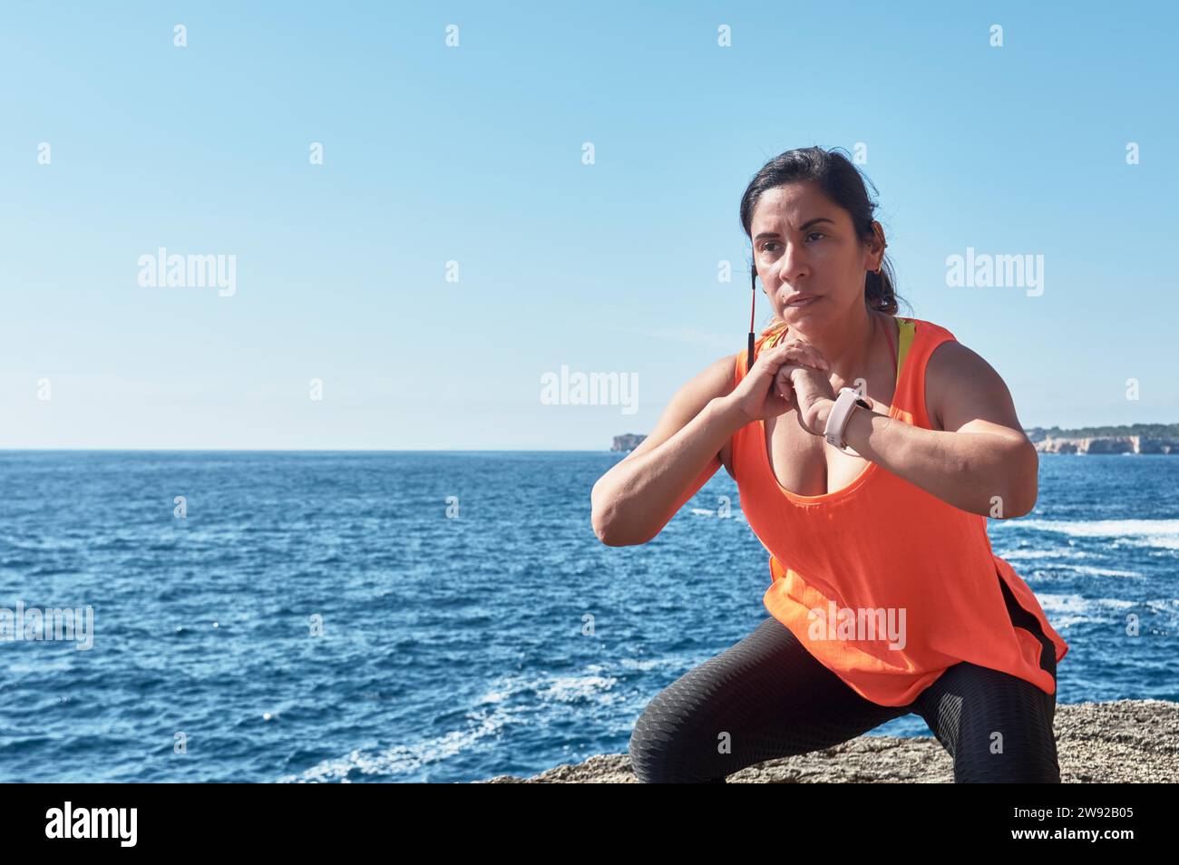 Frau in aktiver Kleidung, die Squats am Meer unter einem klaren blauen Himmel macht Stockfoto