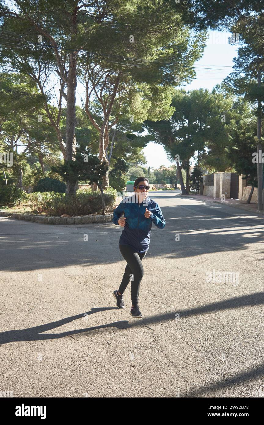 Reife Fitness-Frau, zwischen 30 und 50 Jahre alt, Laufen, unterwegs, umgeben von Pinien, trägt Sportbrille, bluetooth-Kopfhörer, blau Stockfoto