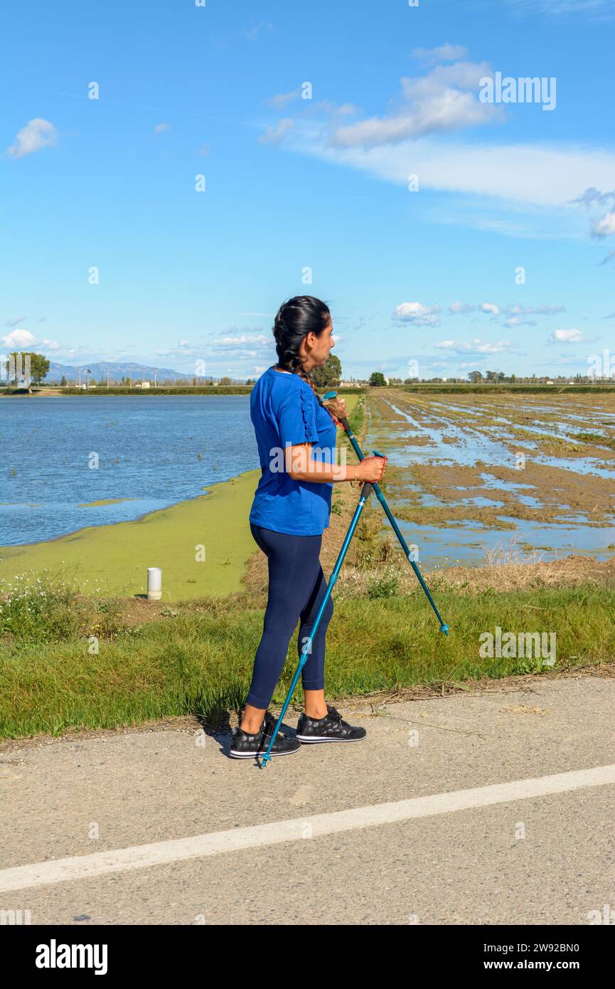 Aktive Person mit Wanderstöcken neben einem Gewässer an einem sonnigen Tag, hispanische Latina-Frau, die mit Trekkingstöcken im natürlichen Ebro-Delta spaziert Stockfoto
