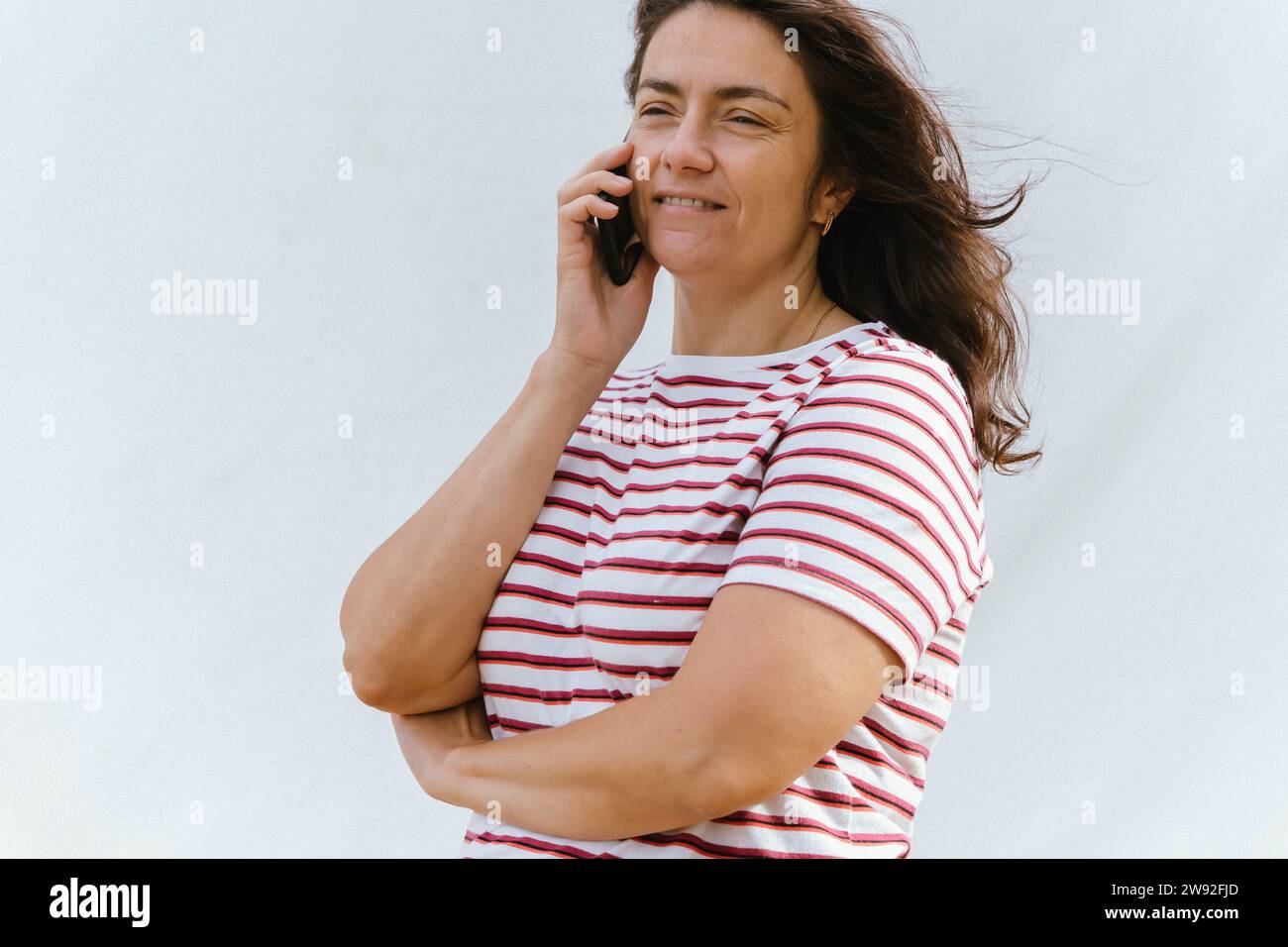 Weiße Frau mittleren Alters mit langen Haaren, die sich auf ihrem Handy fröhlich unterhält. Ihre gestreifte Bluse und der helle Hintergrund unterstreichen den gesamten pi Stockfoto
