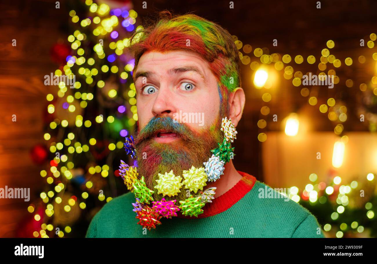 Winterferien. Weihnachts- oder Neujahrsfeier. Überraschter bärtiger Mann mit verziertem Bart und bunten gefärbten Haaren auf weihnachtlichem Hintergrund. Friseur Stockfoto