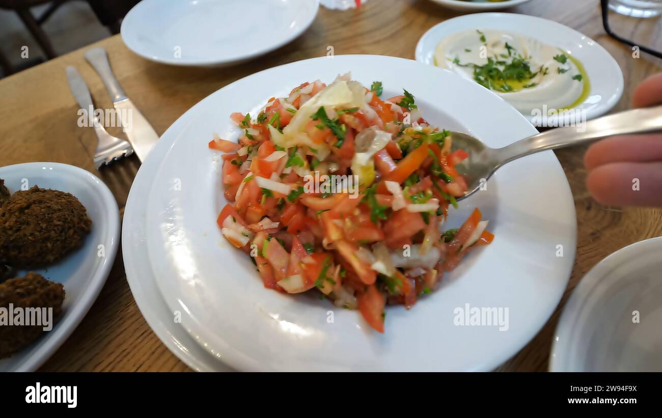 Erfrischender Tomatensalat auf dem Tisch und verführerische Beilagen. Eine Person, die diese kulinarische Symphonie mit einem Löffel genießt. Stockfoto