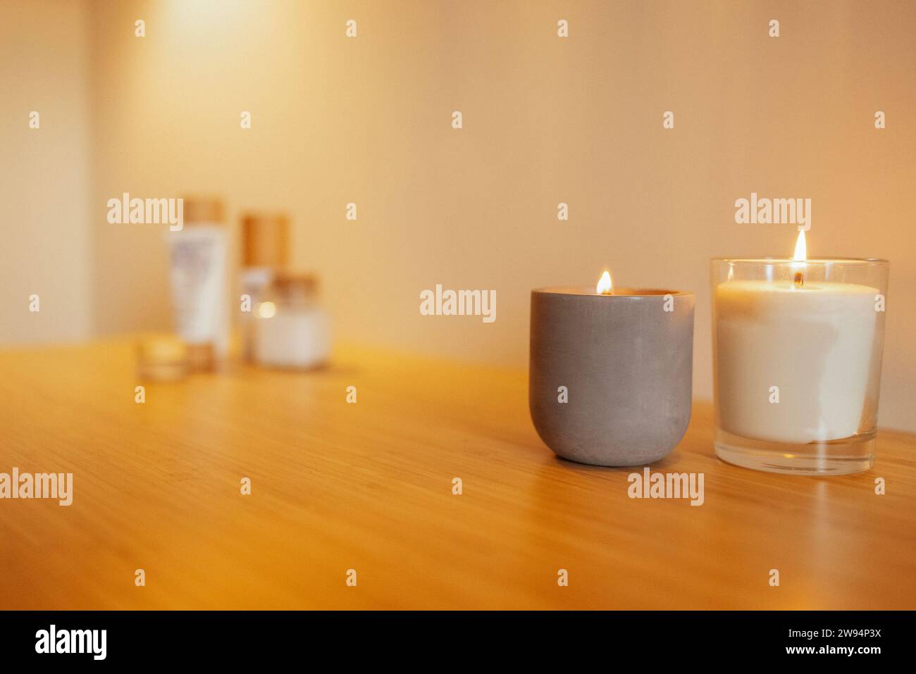 Zwei beleuchtete Kerzen in einem Kerzenhalter aus Keramik und Glas auf einem Holztisch. Eine gemütliche und warme Atmosphäre. Kosmetik- und Schönheitsprodukte im Hintergrund. Stockfoto