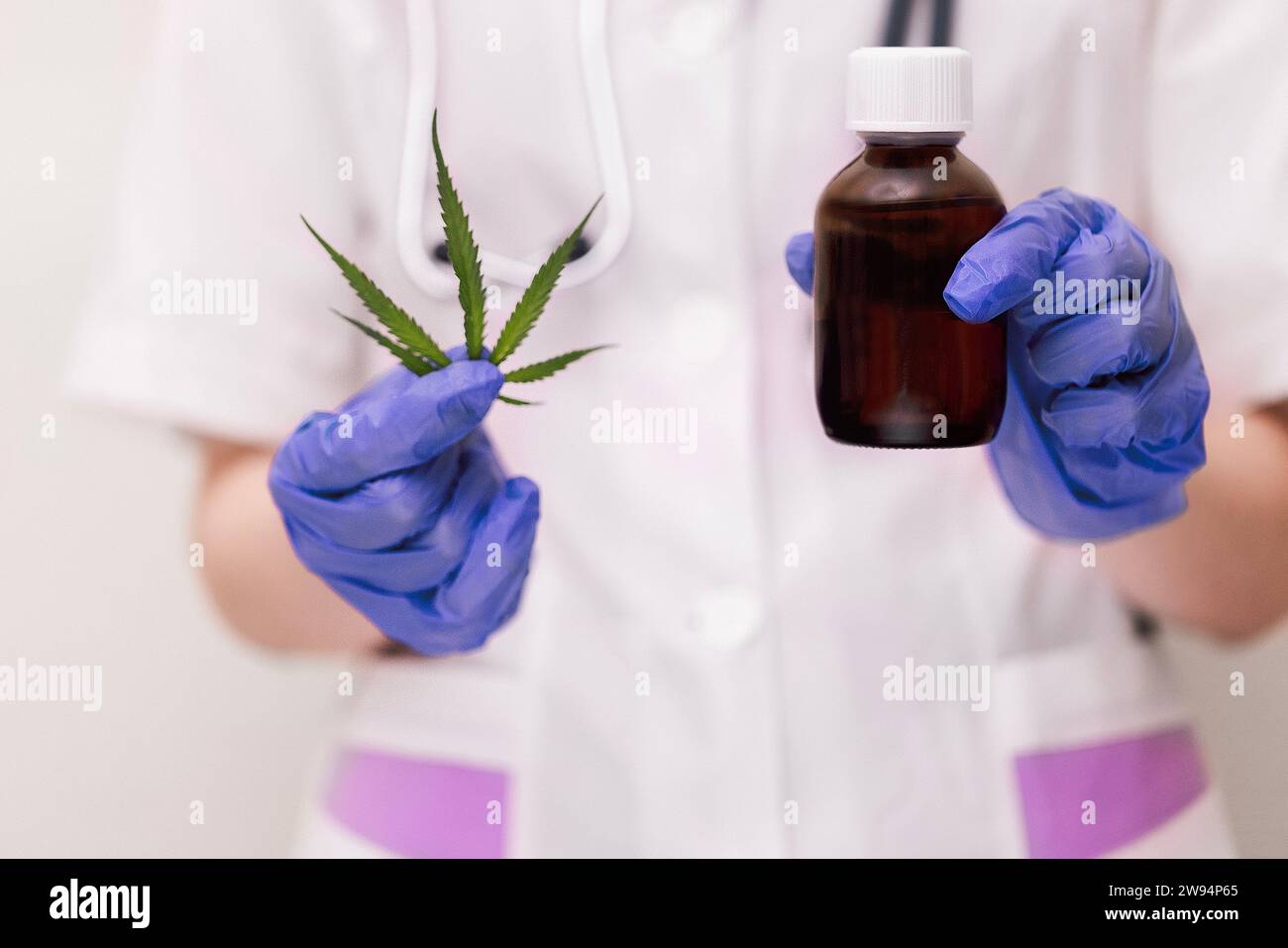 Ein Mediziner in einem Labormantel und blauen Handschuhen hält ein Cannabisblatt und ein Modell einer dunklen Glasflasche Medizin. Hanf-medizinische Eigenschaften Co Stockfoto