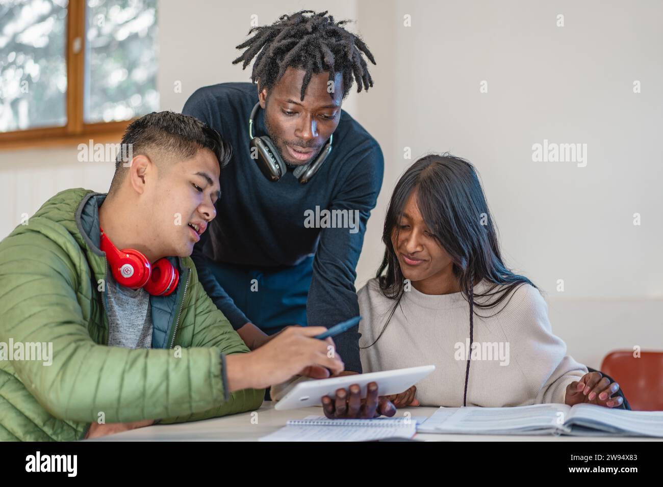 Drei multikulturelle Studenten nehmen an einer interaktiven Lernsitzung mit Notizen und digitalen Geräten Teil und teilen ihr Wissen. Stockfoto
