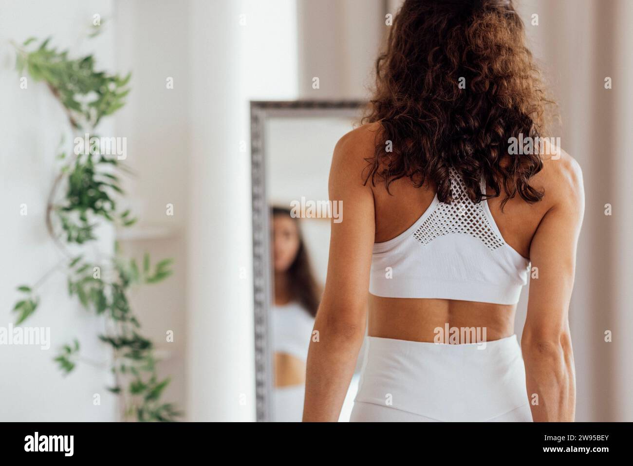 Eine glückliche junge Frau in einem weißen Fitnessanzug genießt morgens das Stretching im Wohnzimmer. Ein lächelndes Mädchen mit lockigen Haaren macht Übungen oder Workouts bei HO Stockfoto