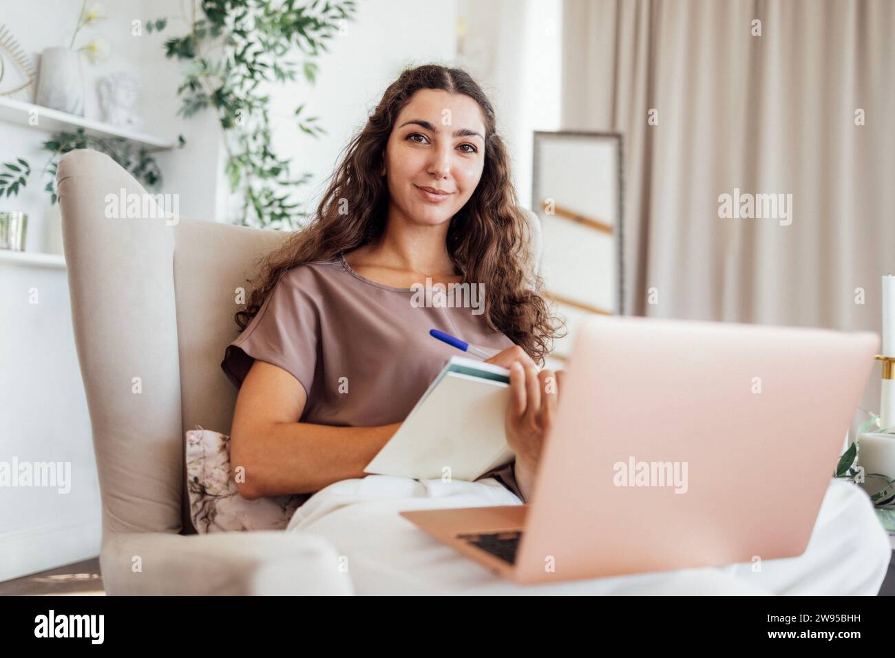 Eine ruhige junge freischaffende Frau in lässiger Kleidung sitzt in einem Sessel mit Notebook und Laptop im Schlafzimmer. Ein charmantes Mädchen mit lockigen Haaren arbeitet Stockfoto