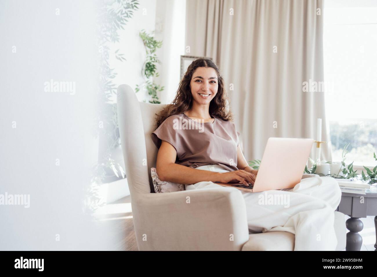 Eine ruhige junge freischaffende Frau in lässiger Kleidung sitzt in einem Sessel mit Notebook und Laptop im Schlafzimmer. Ein charmantes Mädchen mit lockigen Haaren arbeitet Stockfoto