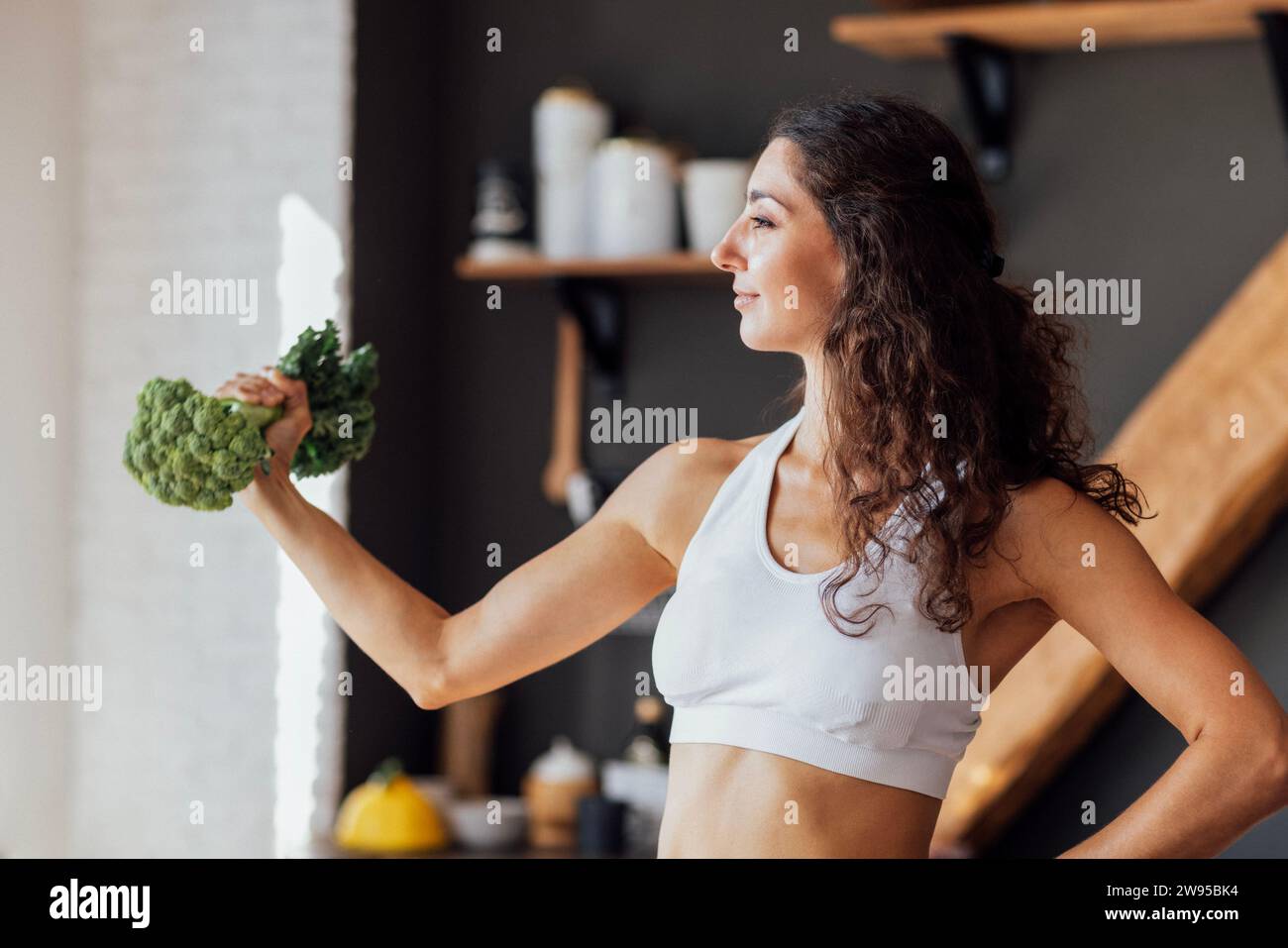 Ein attraktives, lächelndes veganes Mädchen in einem weißen Oberteil beißt frischen Brokkoli. Eine lockige junge Frau lacht und isst Kohl zu Hause. Modern und gemütlich Stockfoto