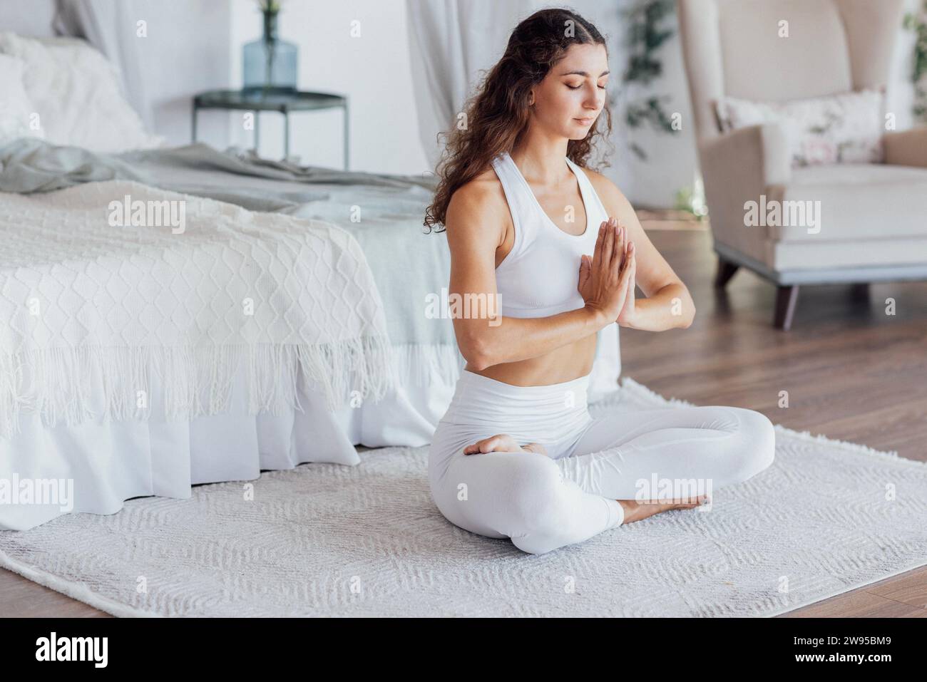 Eine ruhige junge Frau im weißen Fitnessanzug sitzt in Lotusposition und macht morgens im Schlafzimmer Yoga. Ein charmantes Mädchen mit lockigen Haaren genießt s Stockfoto