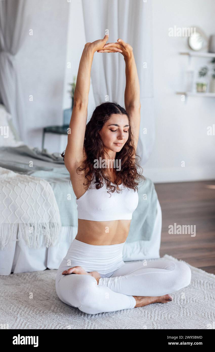Eine ruhige junge Frau im weißen Fitnessanzug sitzt in Lotusposition und macht morgens im Schlafzimmer Yoga. Ein charmantes Mädchen mit lockigen Haaren genießt s Stockfoto