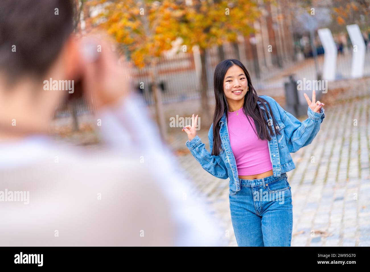 Chinesische junge Frau, die den Sieg mit Gesten angibt, während ein Freund ein Foto auf der Straße mit einer Kamera macht Stockfoto