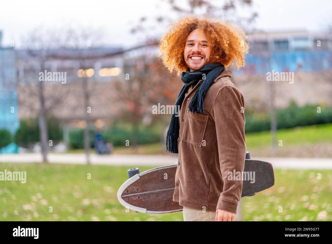 Porträt eines alternativen Mannes mit Skateboard, der in die Kamera lächelt und auf einem Park steht Stockfoto