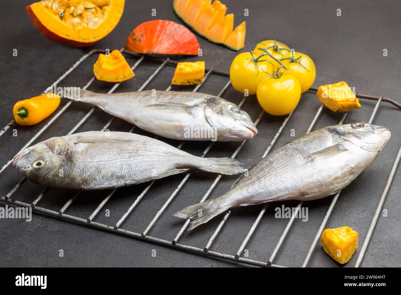Drei rohe dorado-Fische auf dem Grill. Gelbe Tomaten, Paprika und Kürbisscheiben. Schwarzer Hintergrund. Draufsicht Stockfoto