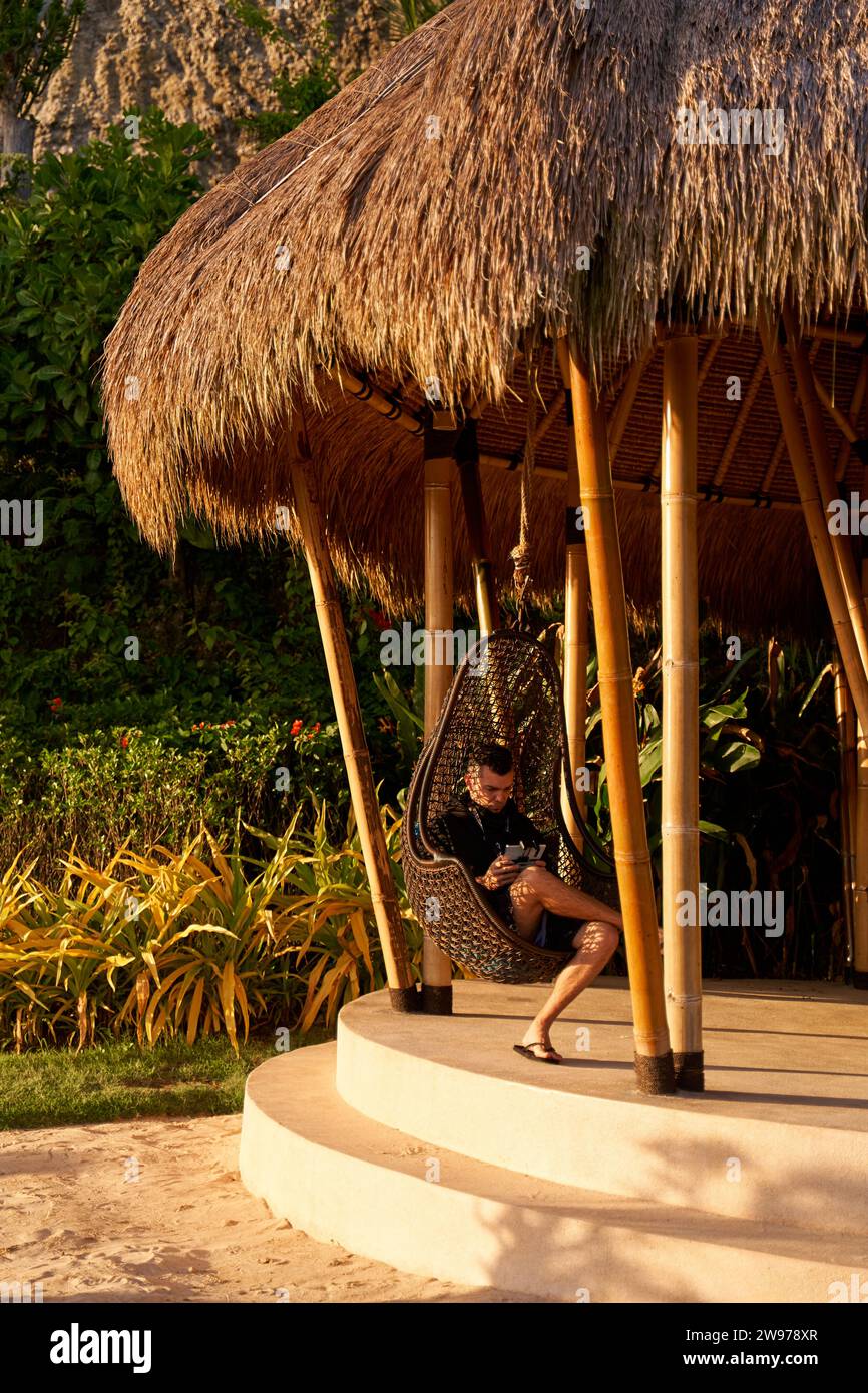 Ein Mann sitzt in einem Schaukelei in einer Stroh-Pergola und genießt den Sonnenuntergang Stockfoto