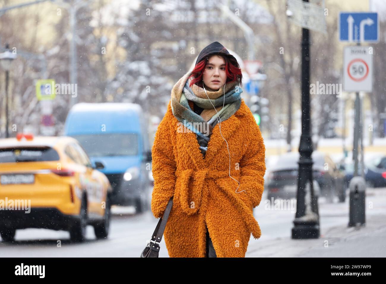Junge Frau im orangefarbenen Mantel, die im Schnee auf der Straße läuft. Winter in einer Stadt, kaltes Wetter Stockfoto