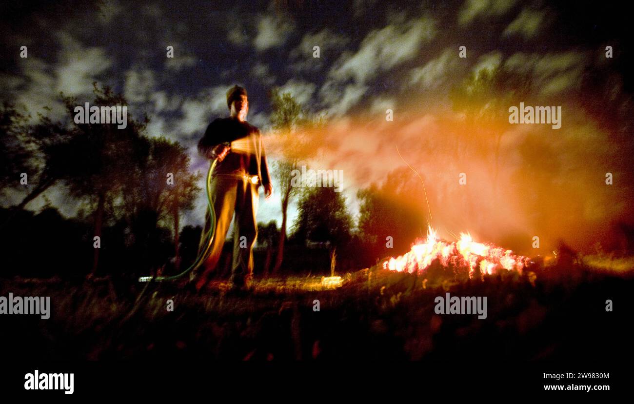 Ein Mann sprüht nachts Wasser auf ein kleines offenes Feuer. (Verschwommenes Bild). Stockfoto