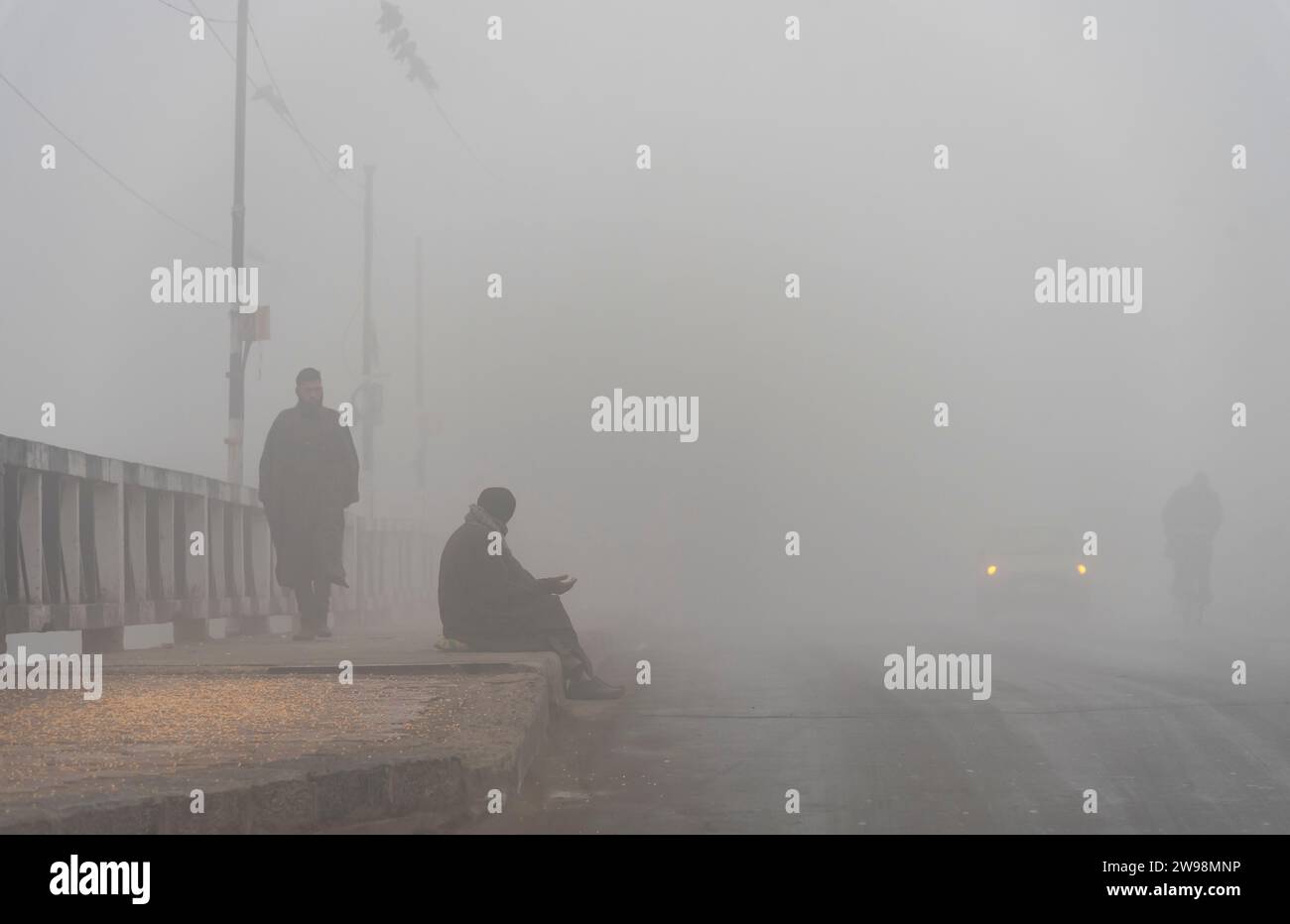 Srinagar, Indien. Dezember 2023. Ein Mann sucht Almosen, während er bei dichtem Nebel in Srinagar auf einem Fußweg sitzt. Intensive Kälte gepaart mit dichtem Nebel und Dunst störte das normale Leben im Kaschmir-Tal am Montag. Srinagar und andere Teile des Kaschmir-Tals waren von einer dicken Schicht Nebel und Dunst umgeben, die die Sicht einschränkte und Pendlern Unannehmlichkeiten bereitete. Quelle: SOPA Images Limited/Alamy Live News Stockfoto