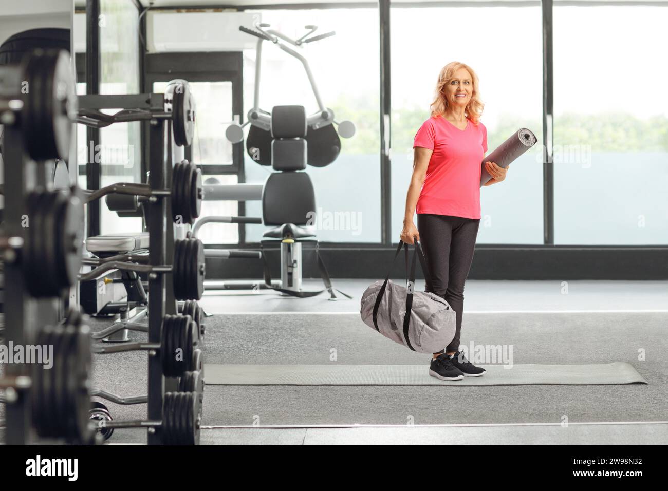 Lächelnde Frau mittleren Alters in Sportbekleidung, die im Fitnessstudio eine Tasche und eine Trainingsmatte hält Stockfoto