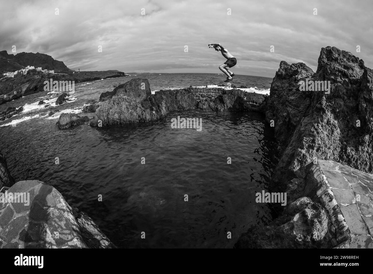 GARACHICO, TENERIFFA, SPANIEN - 28. JULI 2023: Ein Teenager springt ins Wasser. Das felsige Ufer und die natürlichen Pools von El Caleton. Atlantik. Stockfoto