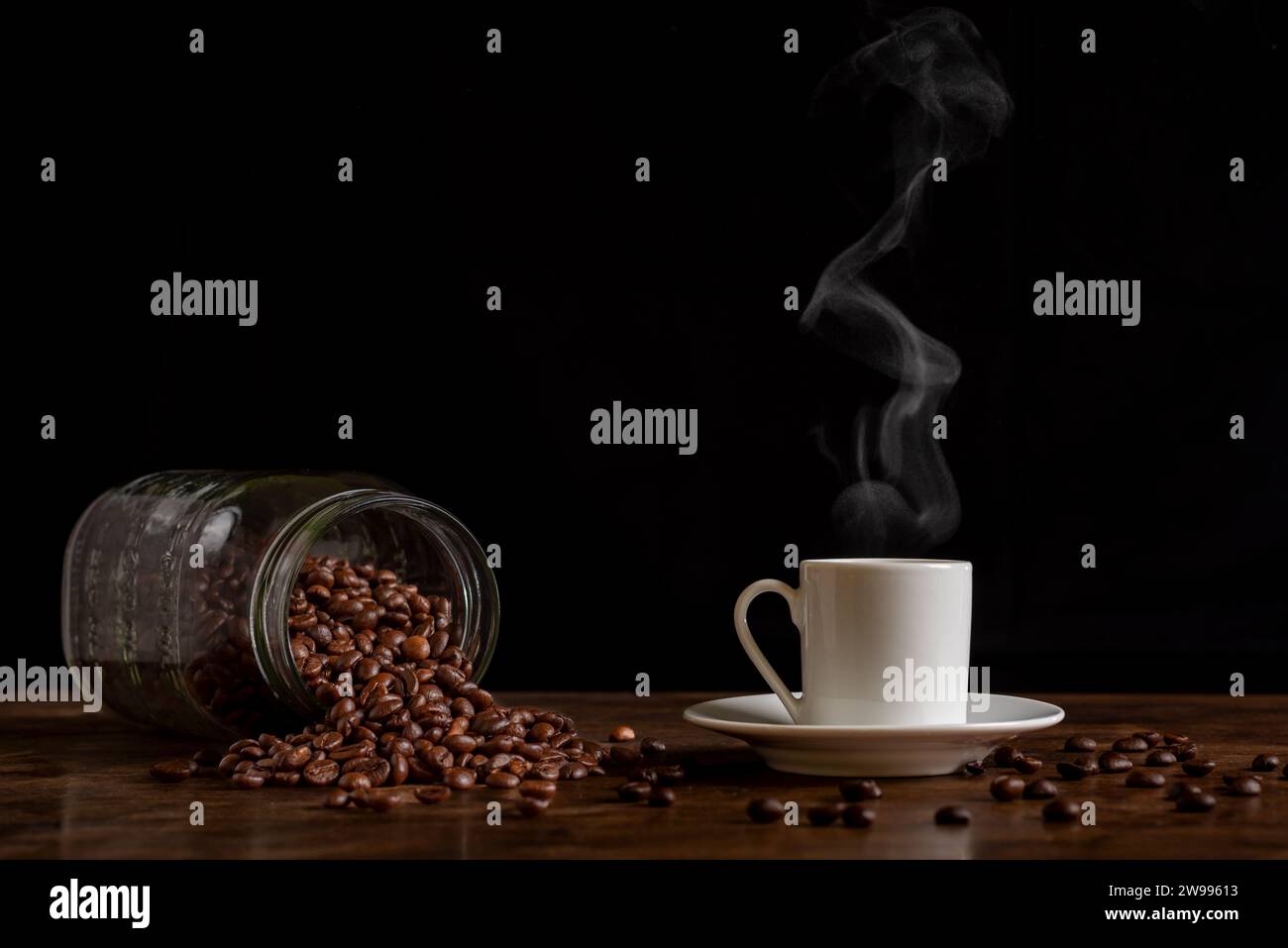 Tasse dampfenden Kaffee neben einem umgedrehten Glas mit Kaffeebohnen auf einem Holztisch mit mehr losen Bohnen, Vorderansicht. Stockfoto