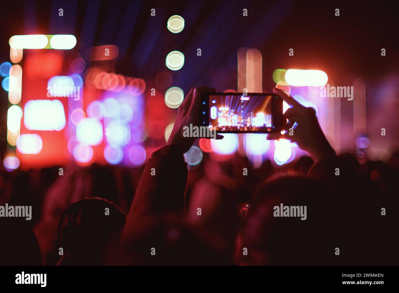 Eine Person nimmt nachts ein Konzert auf dem Skanderberg-Platz mit einem Smartphone auf, mit Bokeh-Bühnenlichtern im Hintergrund Stockfoto