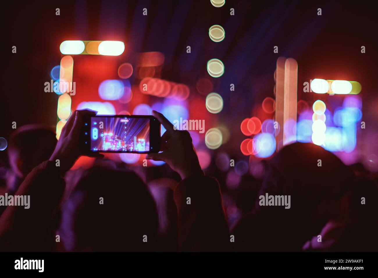 Eine Person nimmt nachts ein Konzert auf dem Skanderberg-Platz mit einem Smartphone auf, mit Bokeh-Bühnenlichtern im Hintergrund Stockfoto