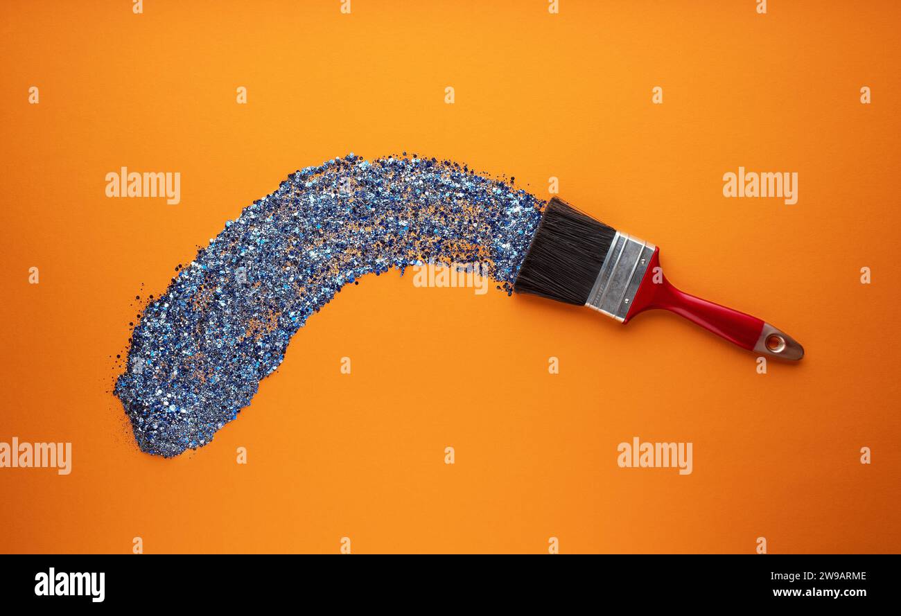 Ein breiter Pinsel zeichnet einen Pinselstrich mit leuchtend blauem Glitzer auf orangem Hintergrund. Stockfoto