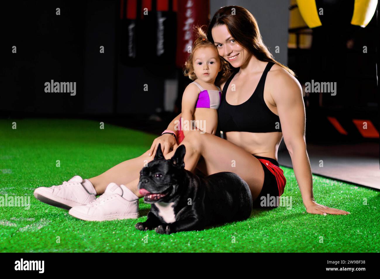 Charmante Sportmutter trainiert im Fitnessstudio mit ihrer kleinen Tochter und einer französischen Bulldogge. Gemischte Medien Stockfoto