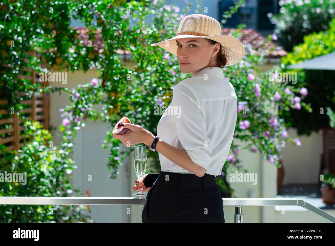 Das charmante Mädchen steht im Garten mit einem Glas Champagner und einem Pfirsich. Das Konzept von Urlaub, Reisen, einem Landhaus. Gemischte Medien Stockfoto