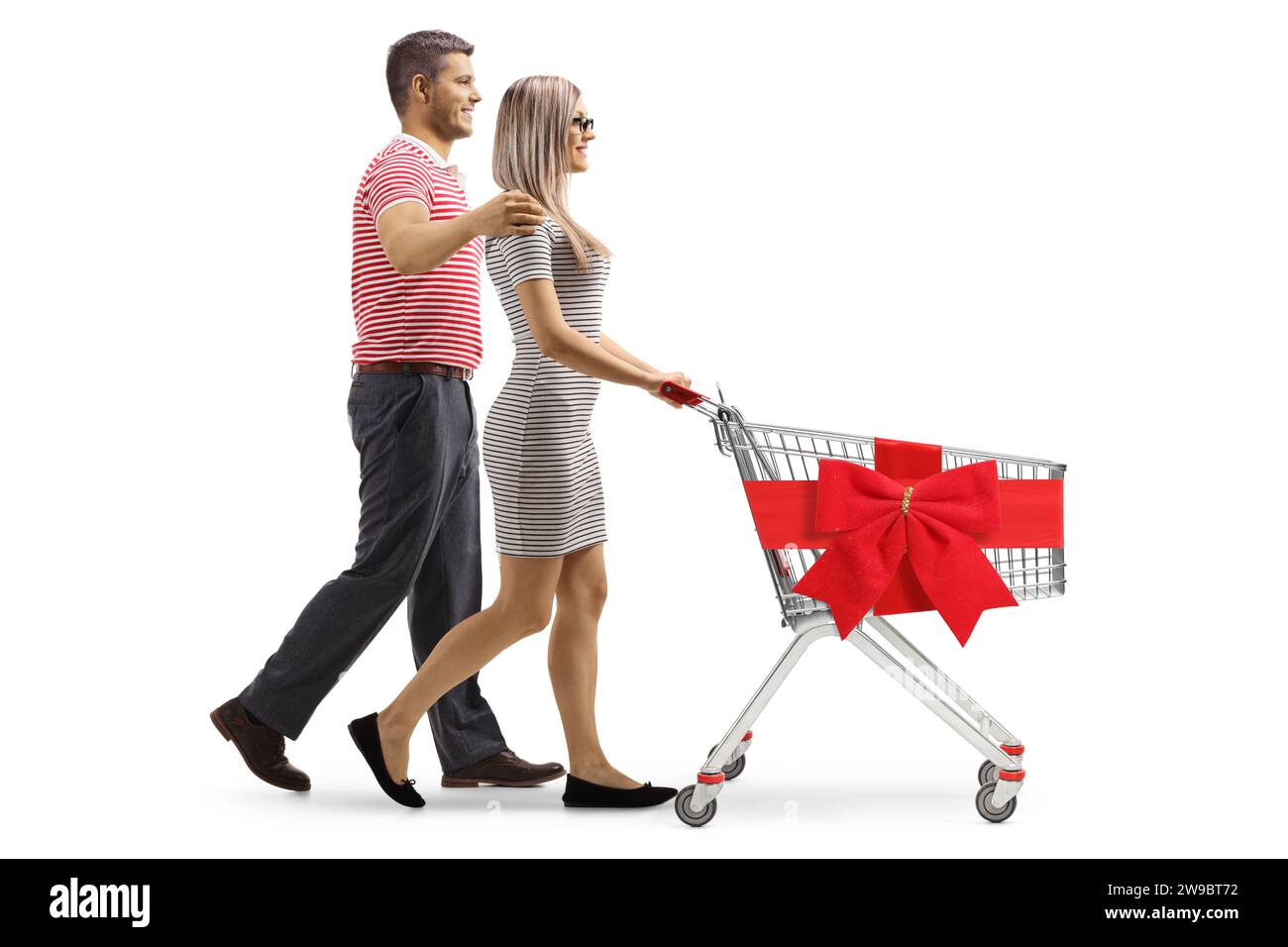 Profilaufnahme eines jungen Paares, das einen Einkaufswagen mit einer roten Schleife auf weißem Hintergrund läuft und schiebt Stockfoto