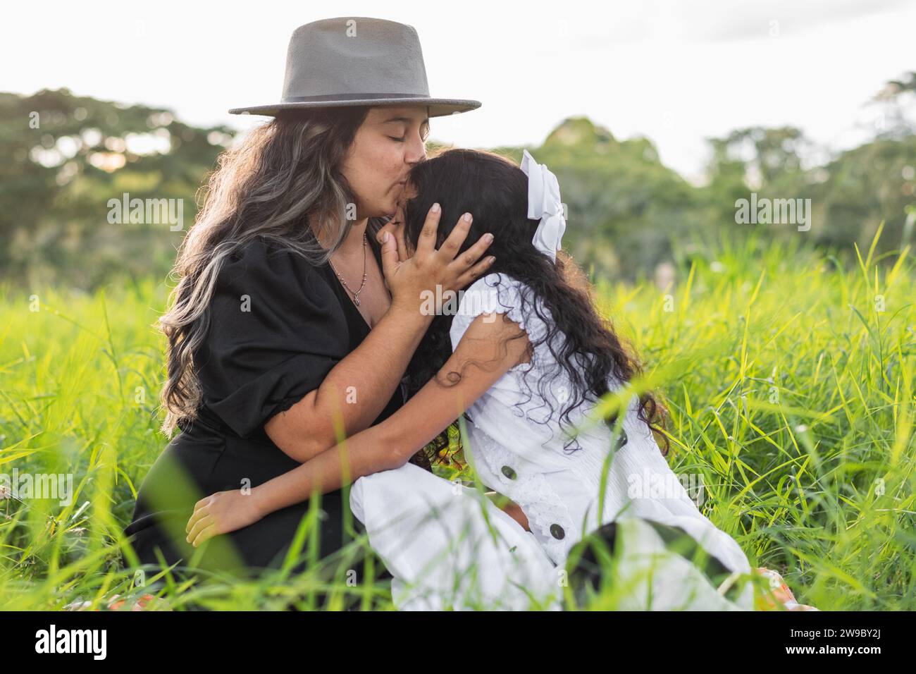 Junge lateinfrau, die ihre kleine Tochter auf die Stirn küsst, während sie auf einem Feld auf hohem Gras sitzt Stockfoto