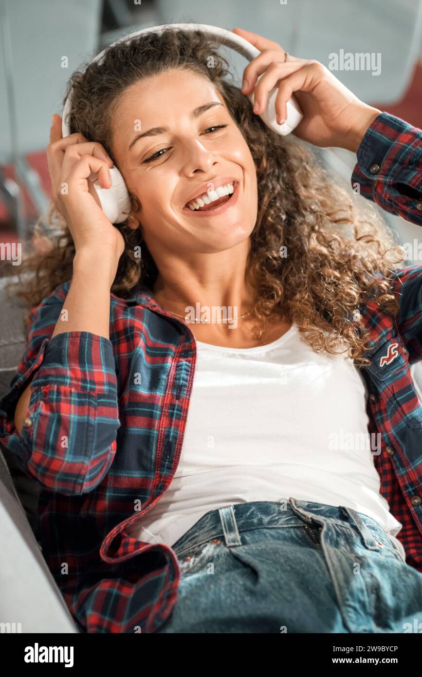 Glückliche junge Frau mit lockigen Haaren in kariertem Hemd, die auf der Couch sitzt und Kopfhörer anpasst, während sie während der Freizeit zu Hause Lieder genießt Stockfoto