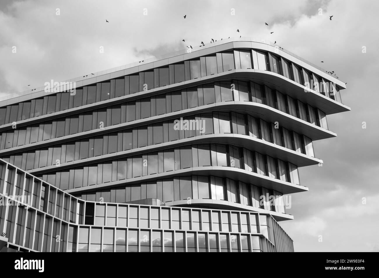 Das auffällige Schwarzweißbild eines geschichteten modernen Gebäudes unter bewölktem Himmel, Vögel im Flug, verstärken die urbane Szene. Stockfoto