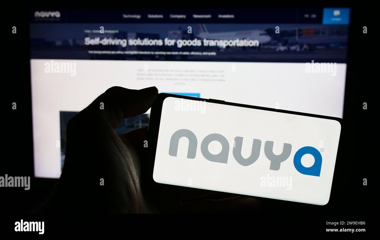 Person, die ein Mobiltelefon mit dem Logo des französischen Autonome Fahrzeugunternehmens Navya S.A. vor der Webseite hält. Schwerpunkt auf dem Display des Telefons. Stockfoto