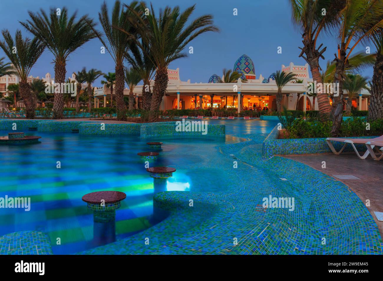 Ein Hotelpool leuchtet mit blauem Mosaiklicht, eingerahmt von Palmensilhouetten und ein lebhaftes Hotelrestaurant in der Ferne. Tropische Oase Bei Nacht. Stockfoto