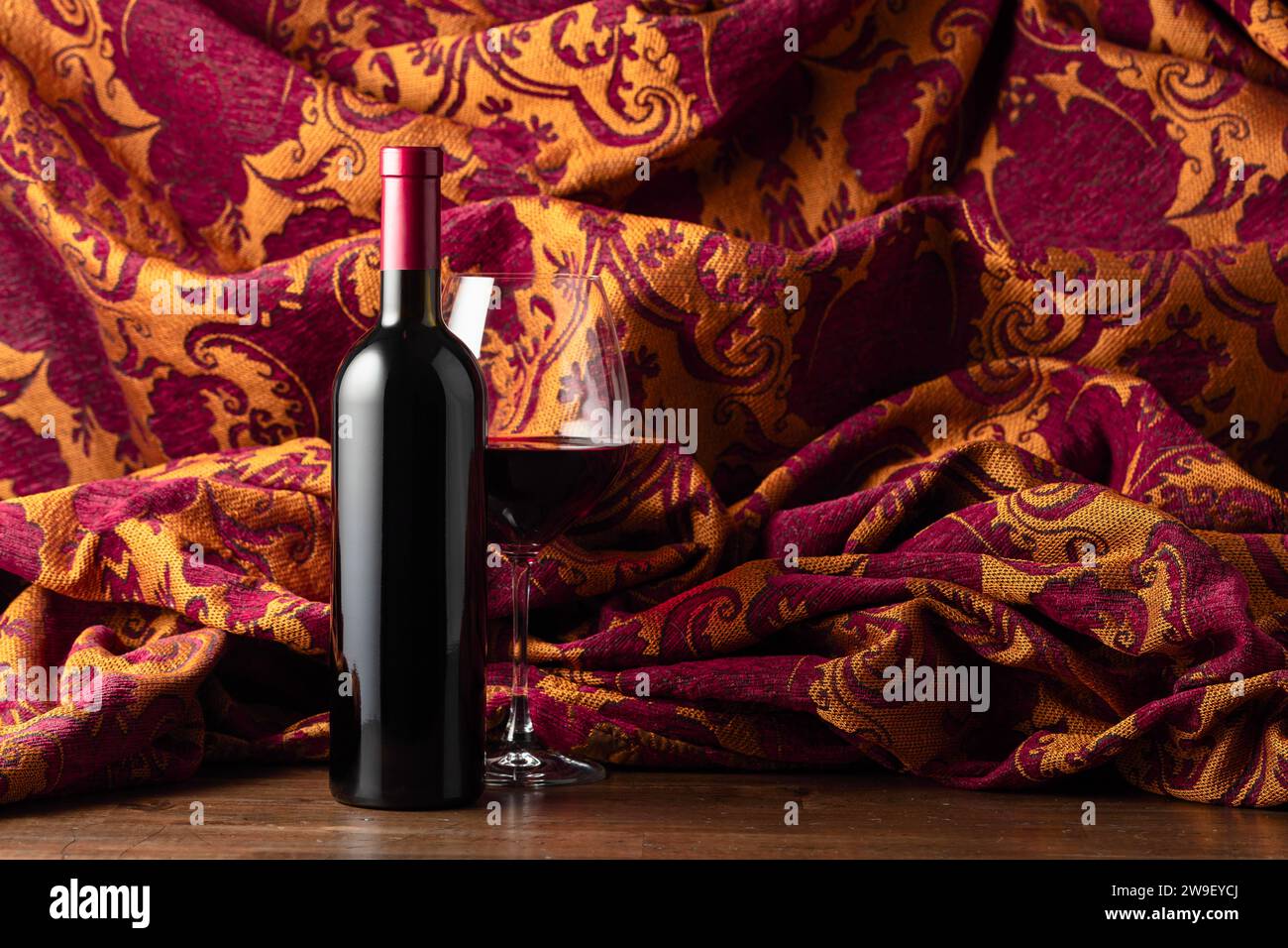 Flasche und Glas Rotwein auf einem alten Holzteller. Im Hintergrund Retro-Wandteppich mit dunkelrotem und goldenem Blumenschmuck. Stockfoto