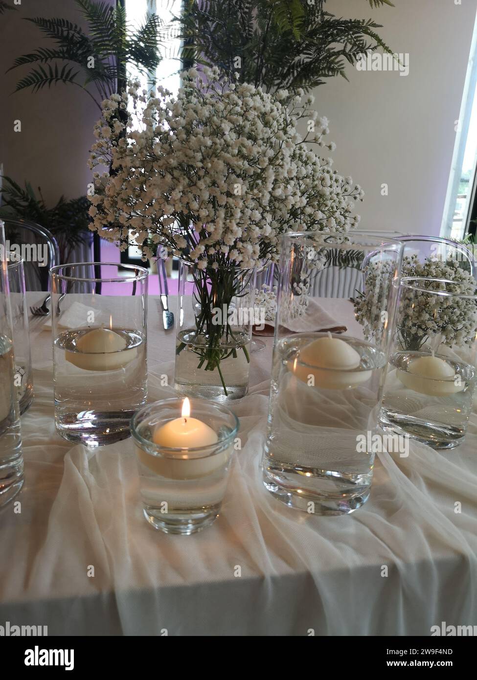Ein Holztisch ist mit mehreren Glasvasen geschmückt, die beleuchtete Kerzen und üppiges Grün beherbergen Stockfoto