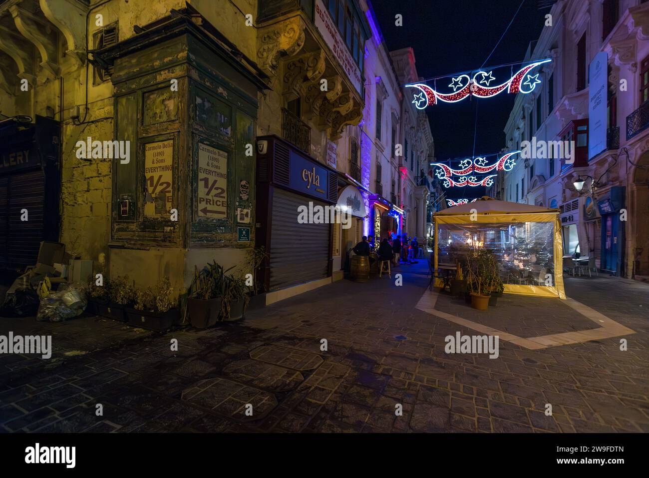 Rote und weiße Lichterketten in Form von Sternen und Halbmonden spannen sich über eine schmale Gasse in der Altstadt von Valletta, Malta, bei Nacht, vor einem beleuchteten Straßencafé unter einem gelben Markisenzelt. Stockfoto