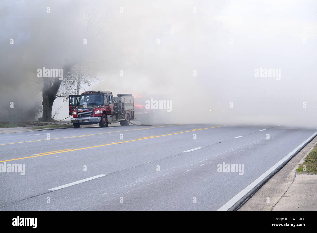Inmitten eines dichten Rauchtuchs eines nahegelegenen Feuers taucht ein leuchtender roter Feuerwehrwagen als Leuchtfeuer der Hoffnung und Aktion auf. Das robuste Fahrzeug steht wie ein Sy Stockfoto