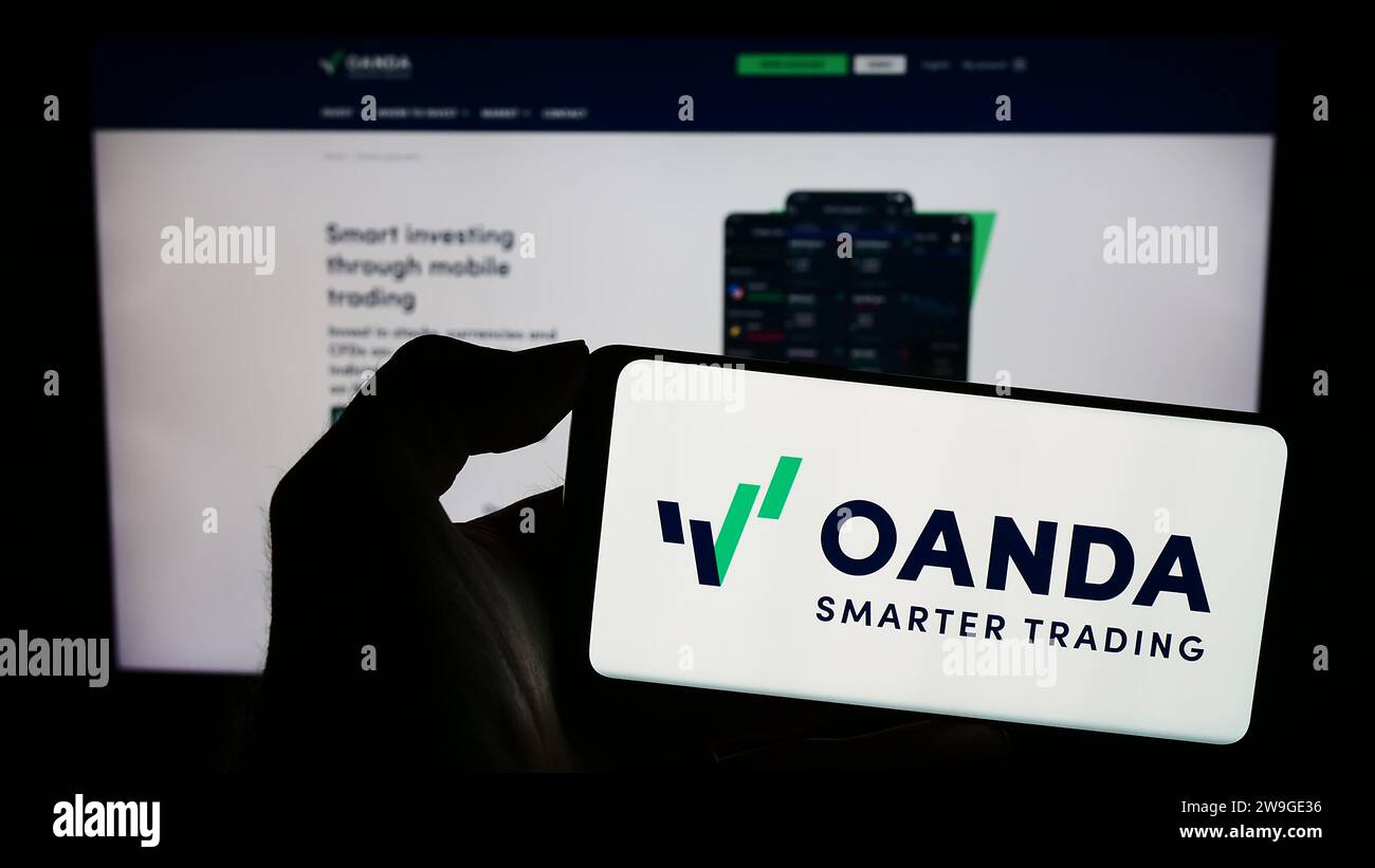 Person, die ein Mobiltelefon mit dem Logo des US-amerikanischen Unternehmens OANDA Business Information Services Inc. Vor der Website des Unternehmens hält. Schwerpunkt auf dem Display des Telefons. Stockfoto