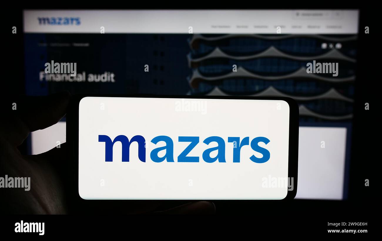 Person, die ein Mobiltelefon mit dem Logo der internationalen Buchhaltungsgesellschaft Mazars Group vor der Business-Webseite hält. Schwerpunkt auf dem Display des Telefons. Stockfoto