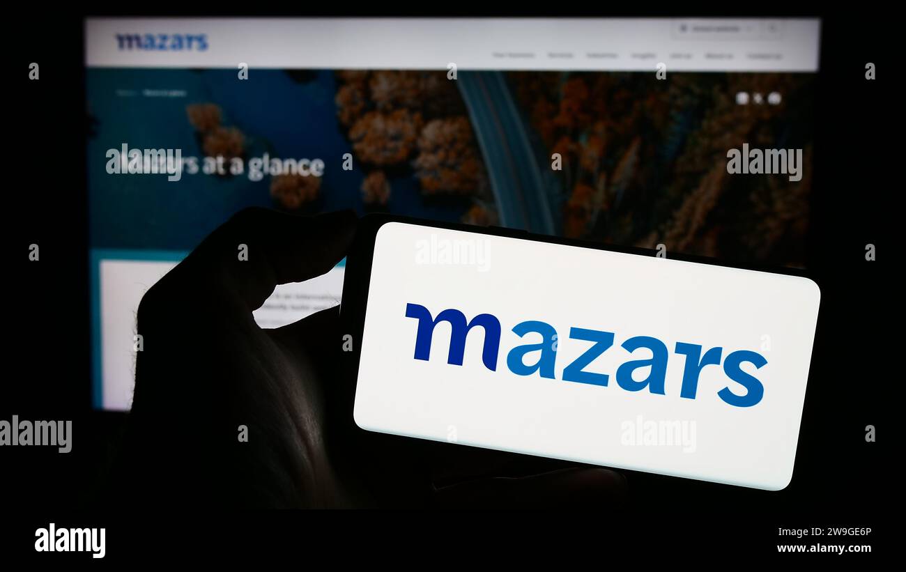 Person, die ein Mobiltelefon mit dem Logo der internationalen Buchhaltungsgesellschaft Mazars Group vor der Webseite hält. Schwerpunkt auf dem Display des Telefons. Stockfoto