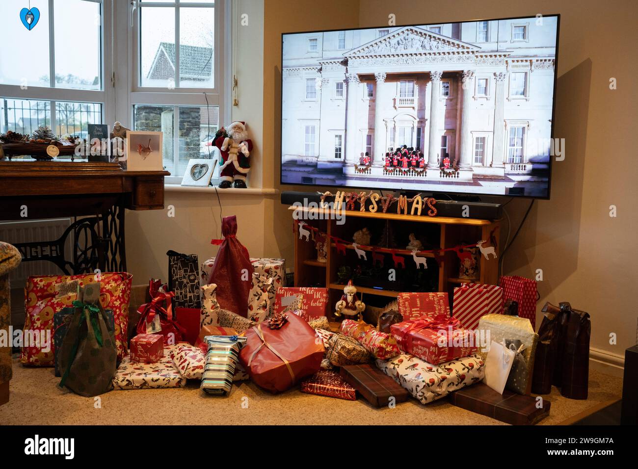 Über den ungeöffneten Weihnachtsgeschenken befinden sich die Eröffnungsszenen, die den Buckingham Palace zeigen, bevor König Charles seine erste Weihnachtssendung als Monarch auf einem Breitbild-TV-Bildschirm im Wohnzimmer eines Familienhauses nach seiner Krönung im Mai, am 25. Dezember 2023, in Nailsea, England gibt. Stockfoto